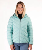 A woman wearing a light mint green hooded puffer jacket with a diagonal quilted pattern, paired with a black top and blue jeans, against a plain white background.