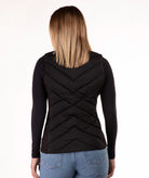 A woman viewed from the back wearing a black quilted puffer vest over a black long-sleeve top and blue jeans, against a plain white studio background.