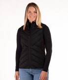 A woman modeling a black zip-front puffer vest over a black long-sleeve top and blue jeans, photographed against a plain white studio background.