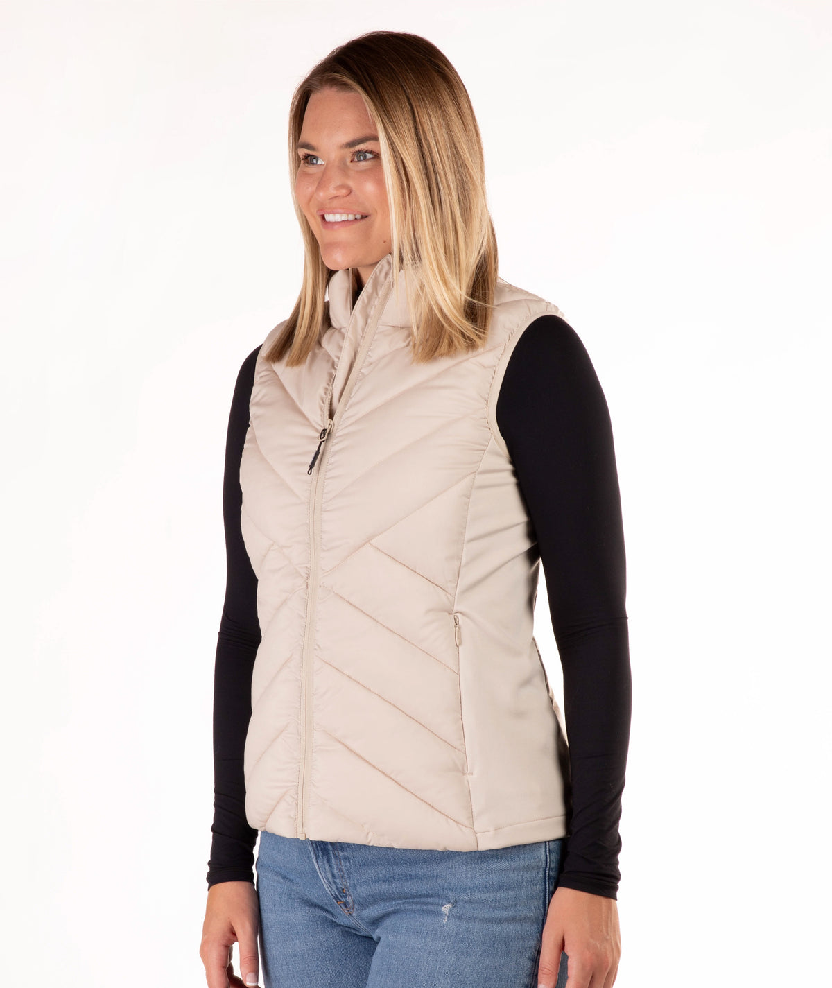 A woman modeling a light beige quilted zip-front vest over a black long-sleeve shirt and blue jeans, photographed against a plain white studio background.