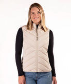 A woman modeling a light beige quilted puffer vest over a black long-sleeve shirt and blue jeans, photographed against a plain white studio background.