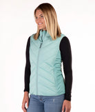 A woman modeling a sleeveless quilted puffer vest in light aqua/mint green, worn over a black long-sleeve shirt and blue jeans, against a plain white studio background.