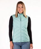 A woman modeling a sleeveless, quilted puffer vest in a light mint green color, worn over a black long-sleeve shirt and blue jeans against a plain white background.