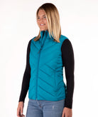 A woman wearing a teal quilted zip-up puffer vest over a black long-sleeve shirt and blue jeans, photographed against a plain white studio background.