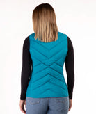 A woman viewed from the back wearing a teal quilted puffer vest over a black long-sleeve top and blue jeans, against a plain white studio background.