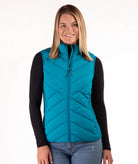 A woman wearing a teal quilted zip-up puffer vest over a black long-sleeve shirt and blue jeans, standing against a plain white studio background.