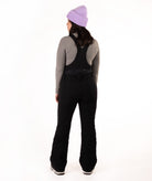 A woman shown from the back wearing black snow bib overalls over a gray long-sleeve top, with a light purple knit beanie, against a plain white studio background.