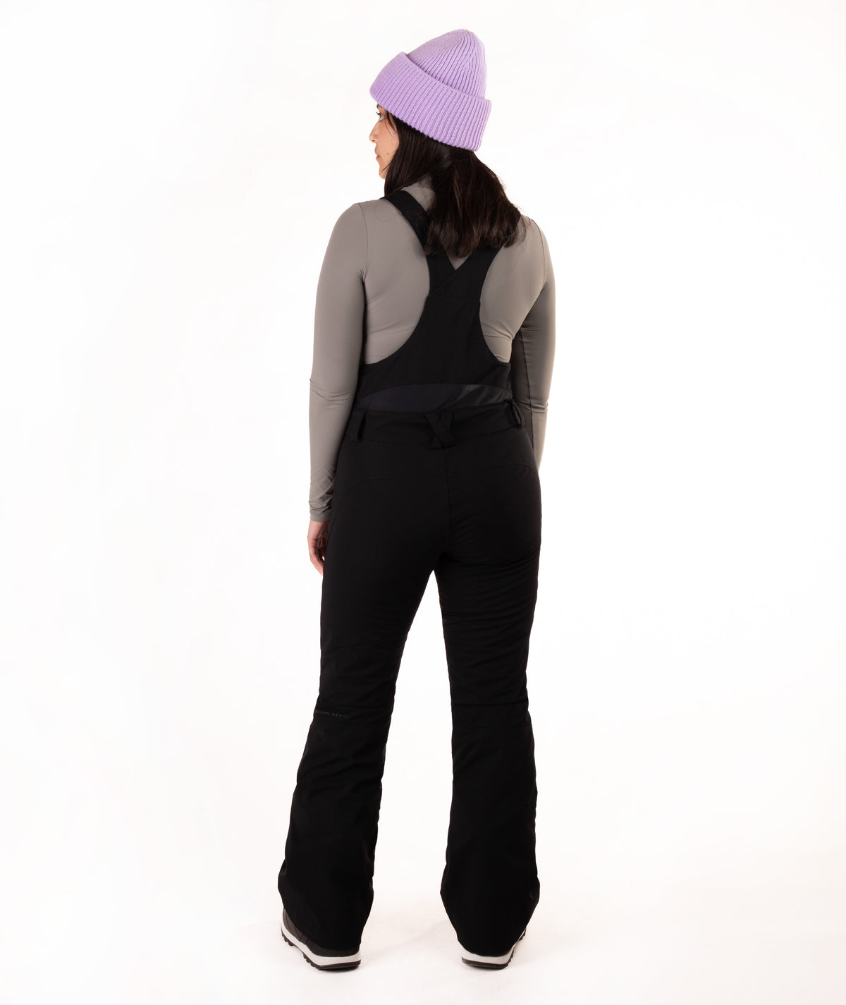 A woman shown from the back wearing black snow bib overalls over a gray long-sleeve top, with a light purple knit beanie, against a plain white studio background.