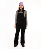 A woman modeling black snow bib overalls over a gray long-sleeve shirt, wearing a light purple knit beanie and black boots, against a solid white studio background.