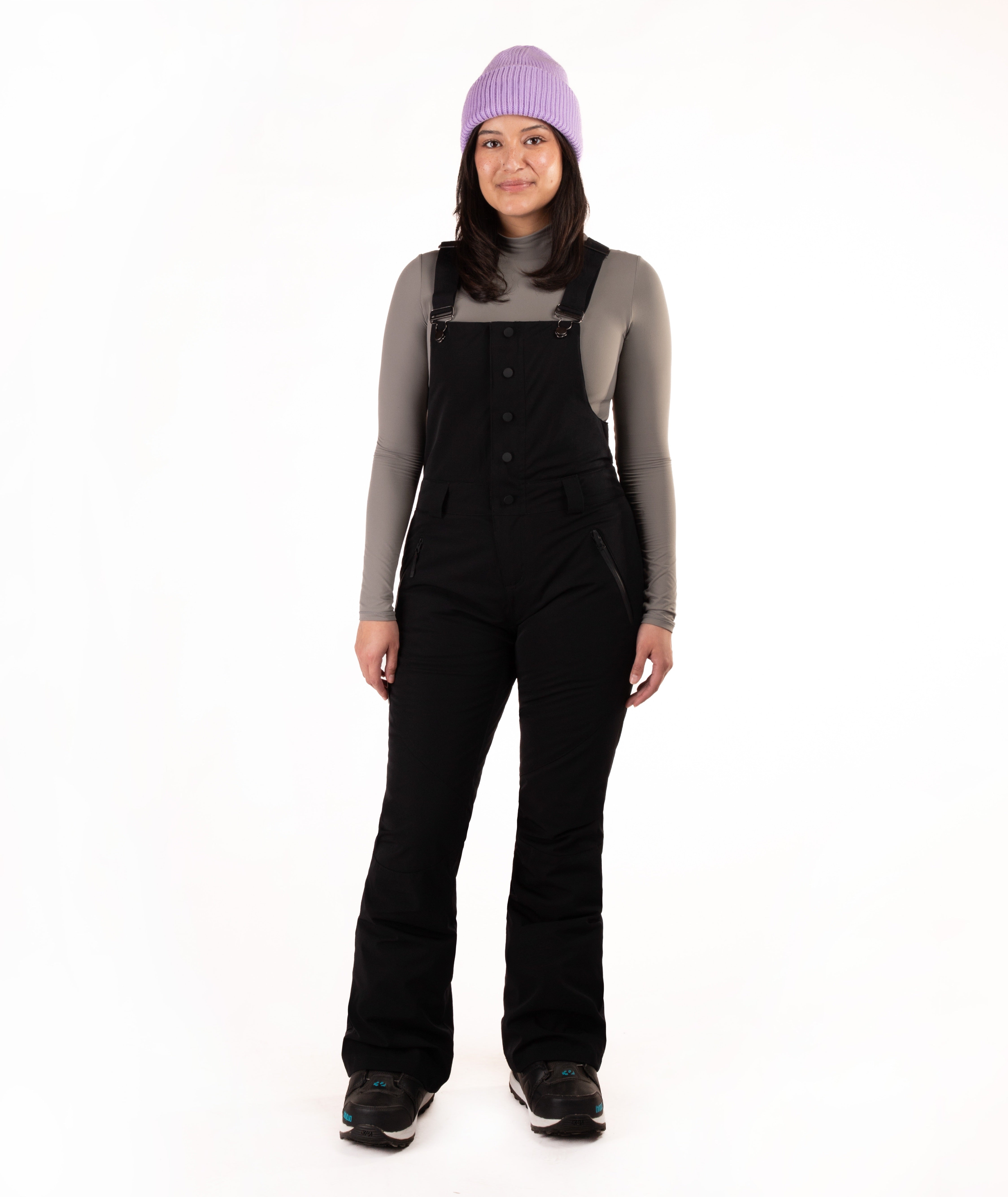 A woman modeling black snow bib overalls over a gray long-sleeve shirt, wearing a light purple knit beanie and black boots, against a solid white studio background.