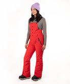 A woman modeling bright red snow bib overalls with black trim and buttons, worn over a gray long-sleeve top, black boots, and a lavender knit beanie against a plain white background.