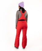 A woman shown from the back wearing bright red ski bib pants over a gray long-sleeve top, with a light purple knit beanie, against a plain white studio background.