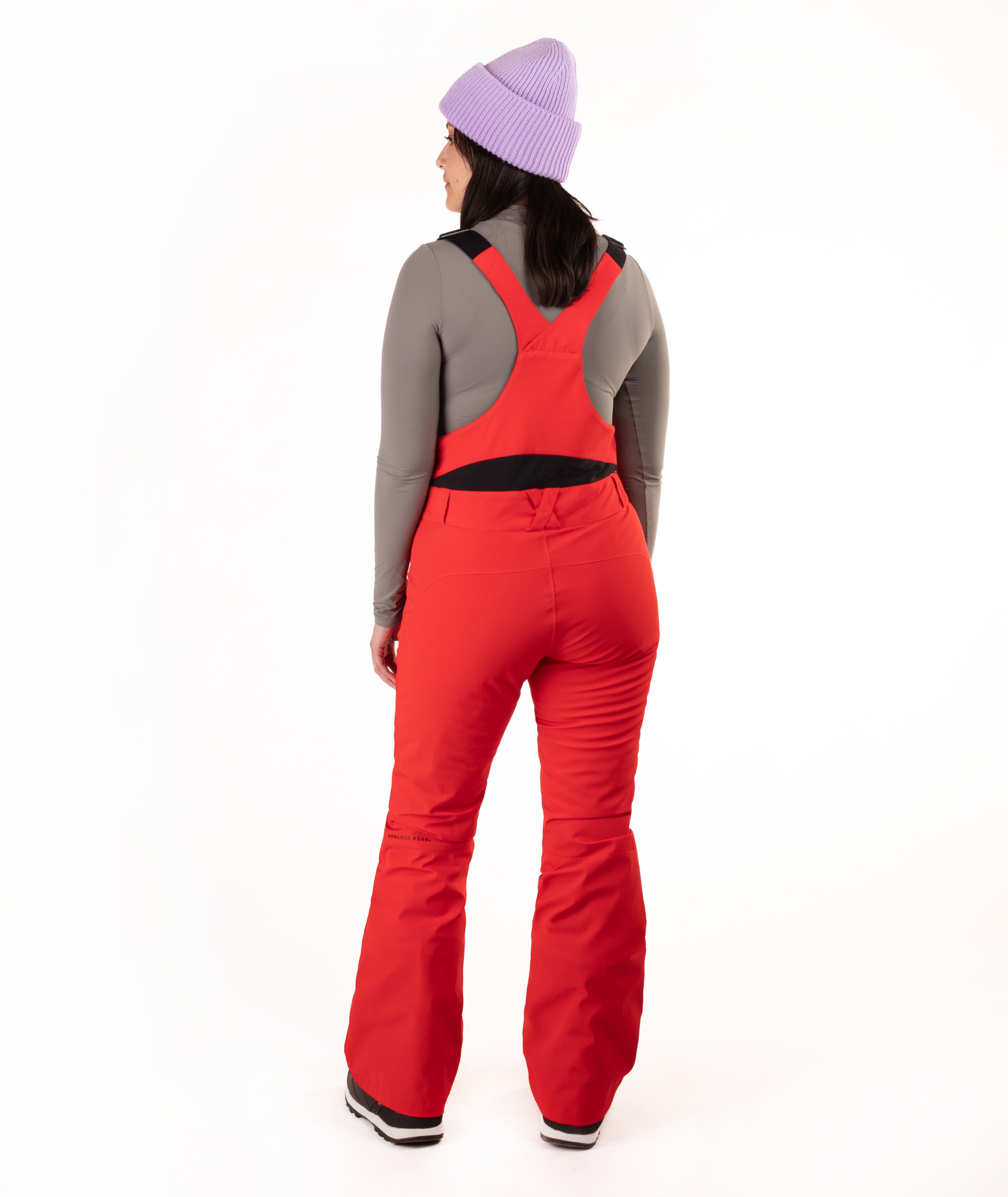 A woman shown from the back wearing bright red ski bib pants over a gray long-sleeve top, with a light purple knit beanie, against a plain white studio background.