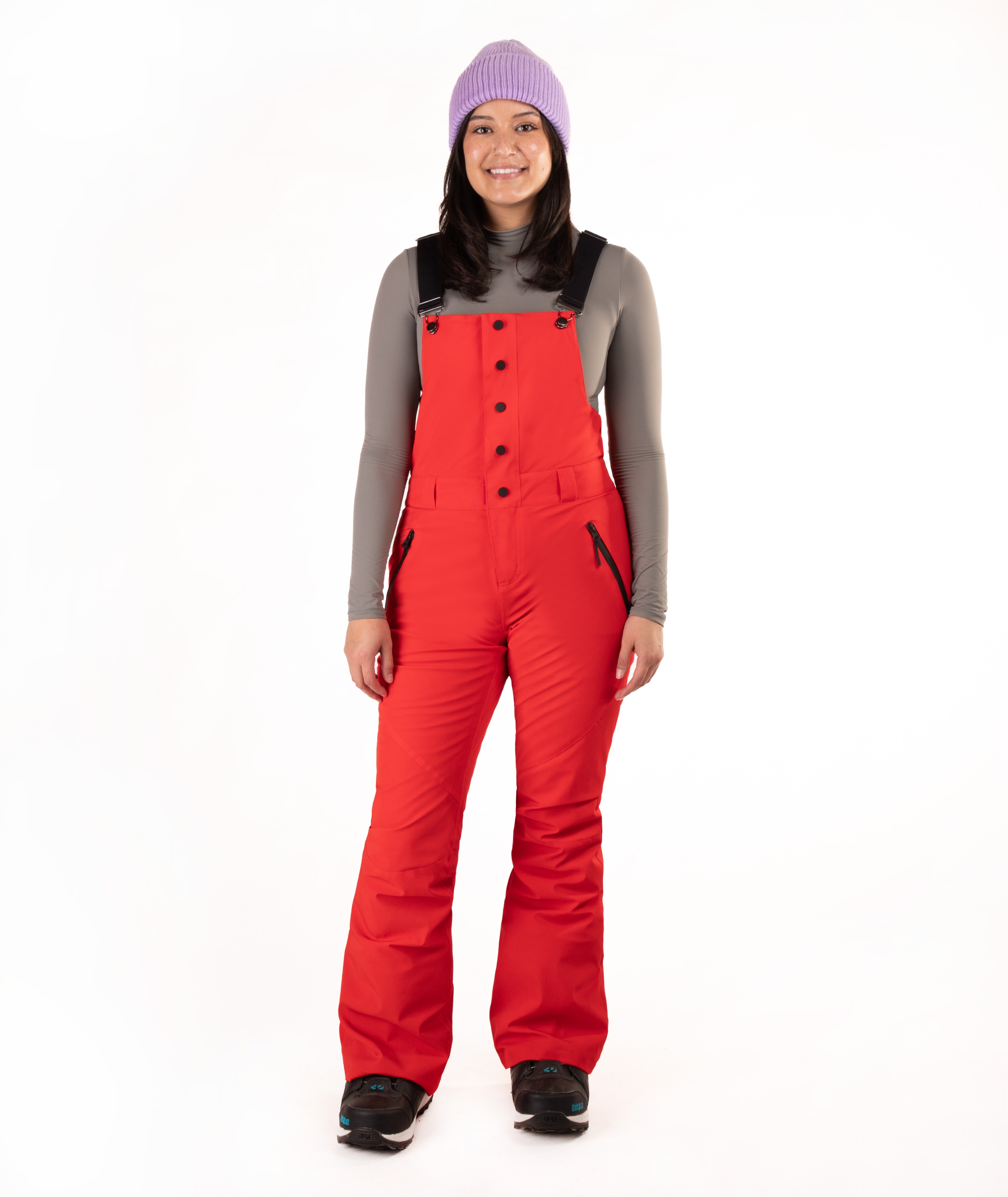 A woman modeling bright red snow bib overalls with black trim and buttons, worn over a gray long-sleeve top, black boots, and a lavender knit beanie against a plain white background.