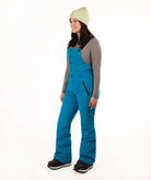 A woman modeling bright teal snow bib overalls with a gray long-sleeve shirt, a light green knit beanie, and black boots, standing against a solid white studio background.
