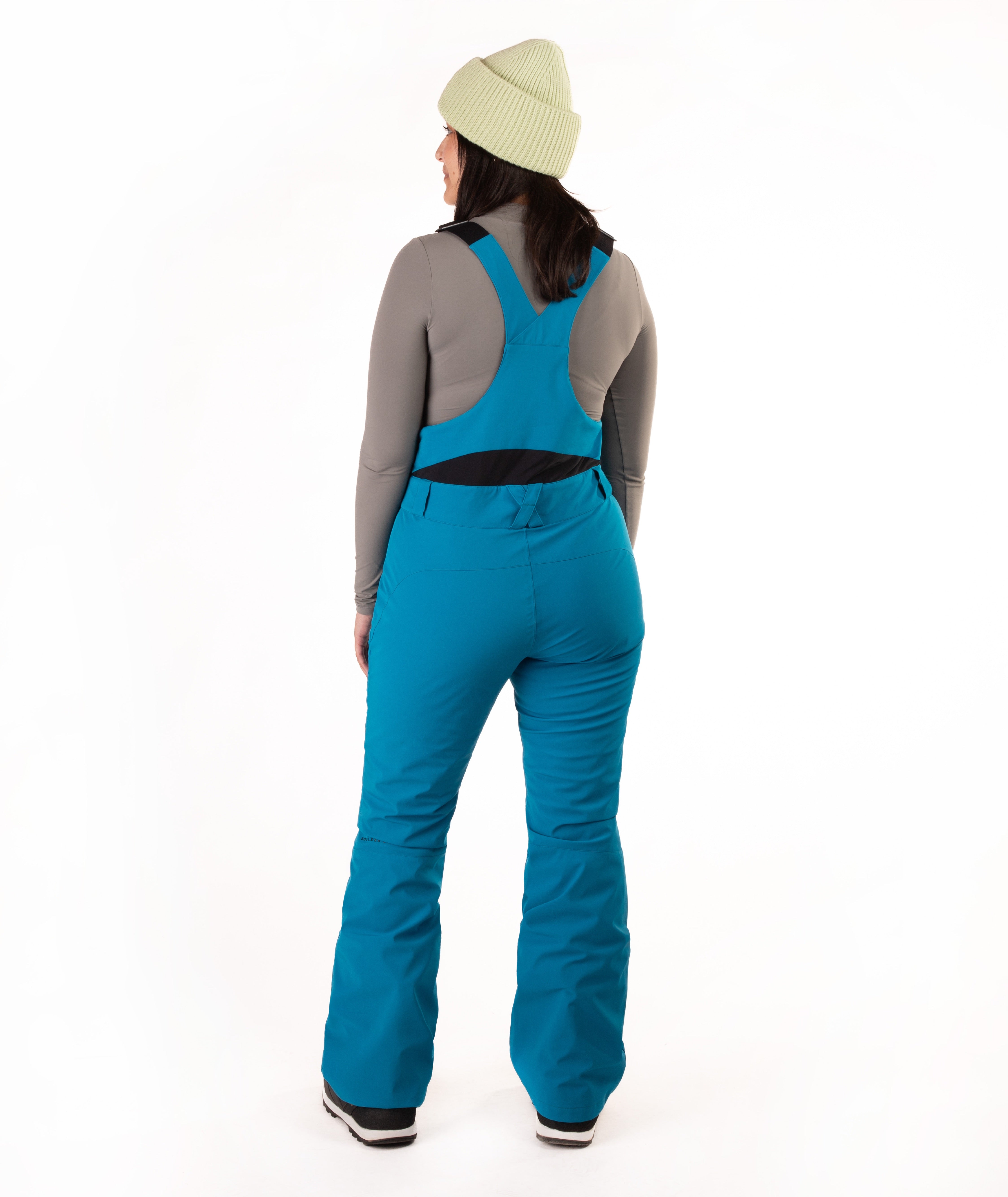 A woman viewed from the back wearing bright teal snow pants over a gray long-sleeve top and a pale green beanie, standing against a plain white studio background.