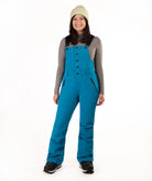 A woman modeling bright teal snow bib overalls with a gray long-sleeve shirt, a light green knit beanie, and black boots, standing against a solid white studio background.