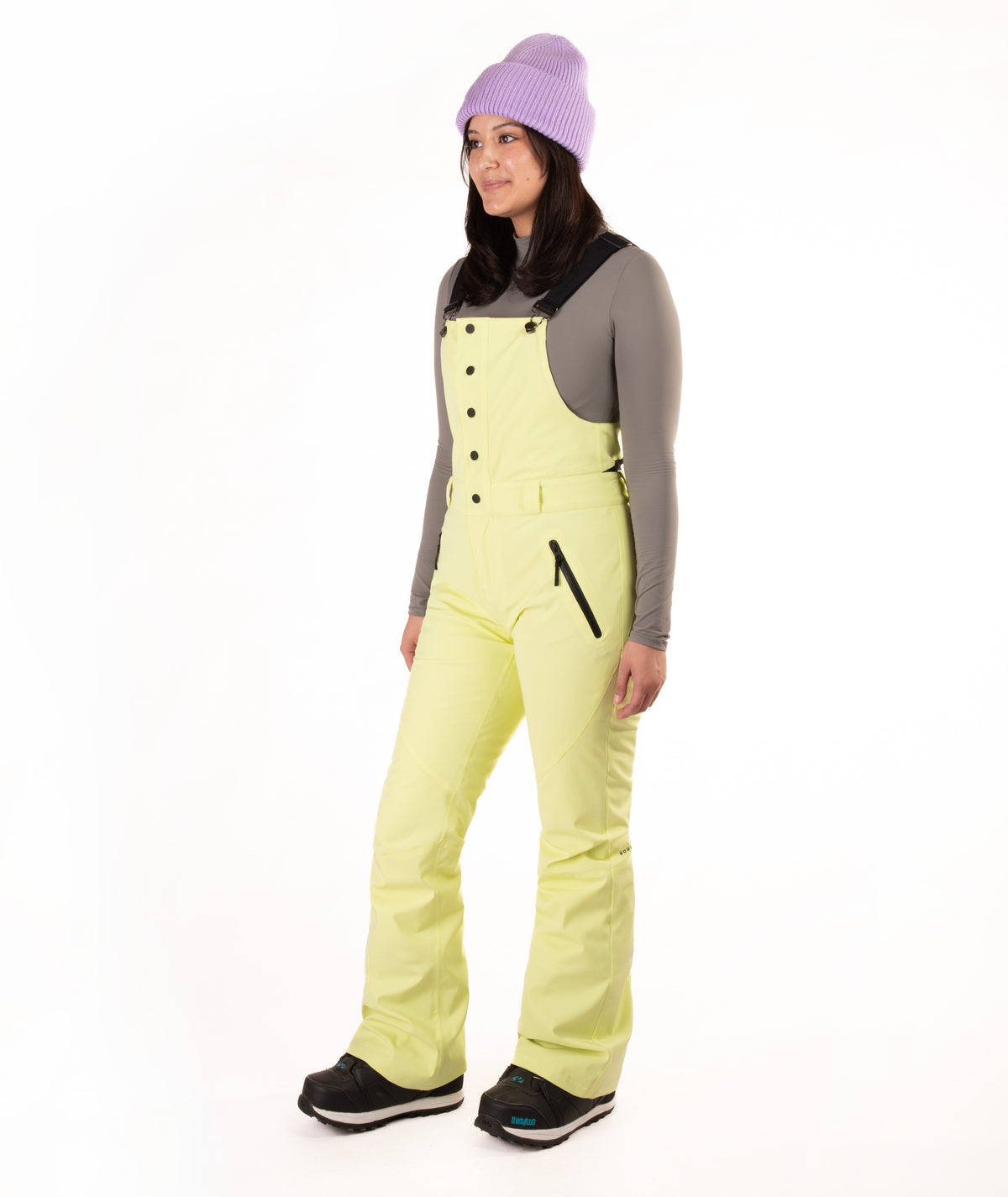 A woman modeling light yellow-green snow bib overalls with black trim and a black button-front bib panel, worn over a gray long-sleeve shirt, black boots, and a lavender knit beanie on a white studio background.