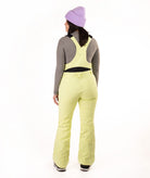 A model shown from the back wearing  light yellow-green ski bibs, a gray long-sleeve top, a purple knit beanie, and black and white boots, set against a plain white studio background.