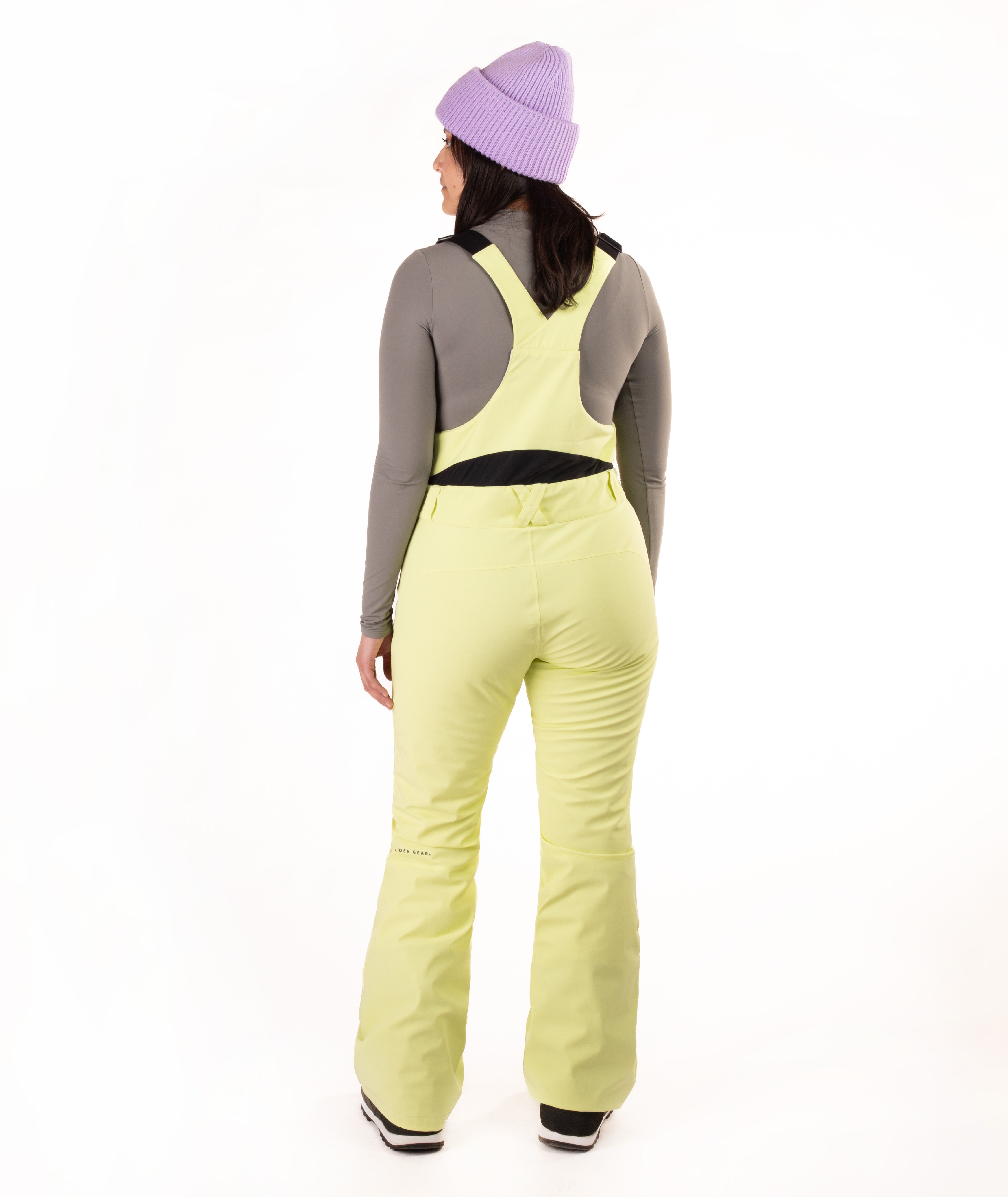 A model shown from the back wearing  light yellow-green ski bibs, a gray long-sleeve top, a purple knit beanie, and black and white boots, set against a plain white studio background.