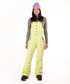 A woman modeling light yellow-green snow bib overalls with black trim and a black button-front bib panel, worn over a gray long-sleeve shirt, black boots, and a lavender knit beanie on a white studio background.