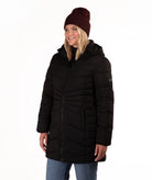 A woman wearing a black hooded puffer jacket, a dark burgundy knit beanie, and blue jeans, photographed against a plain white studio background.