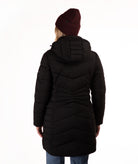 A woman viewed from behind wearing a long black hooded puffer coat, a dark burgundy knit beanie, and blue jeans against a plain white studio background.