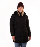A woman wearing a black hooded puffer coat, a dark burgundy knit beanie, and blue jeans, standing against a plain white background.