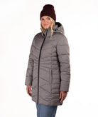 A woman wearing a gray hooded puffer jacket with a black front zipper, paired with a burgundy knit beanie and blue jeans, standing against a plain white background.