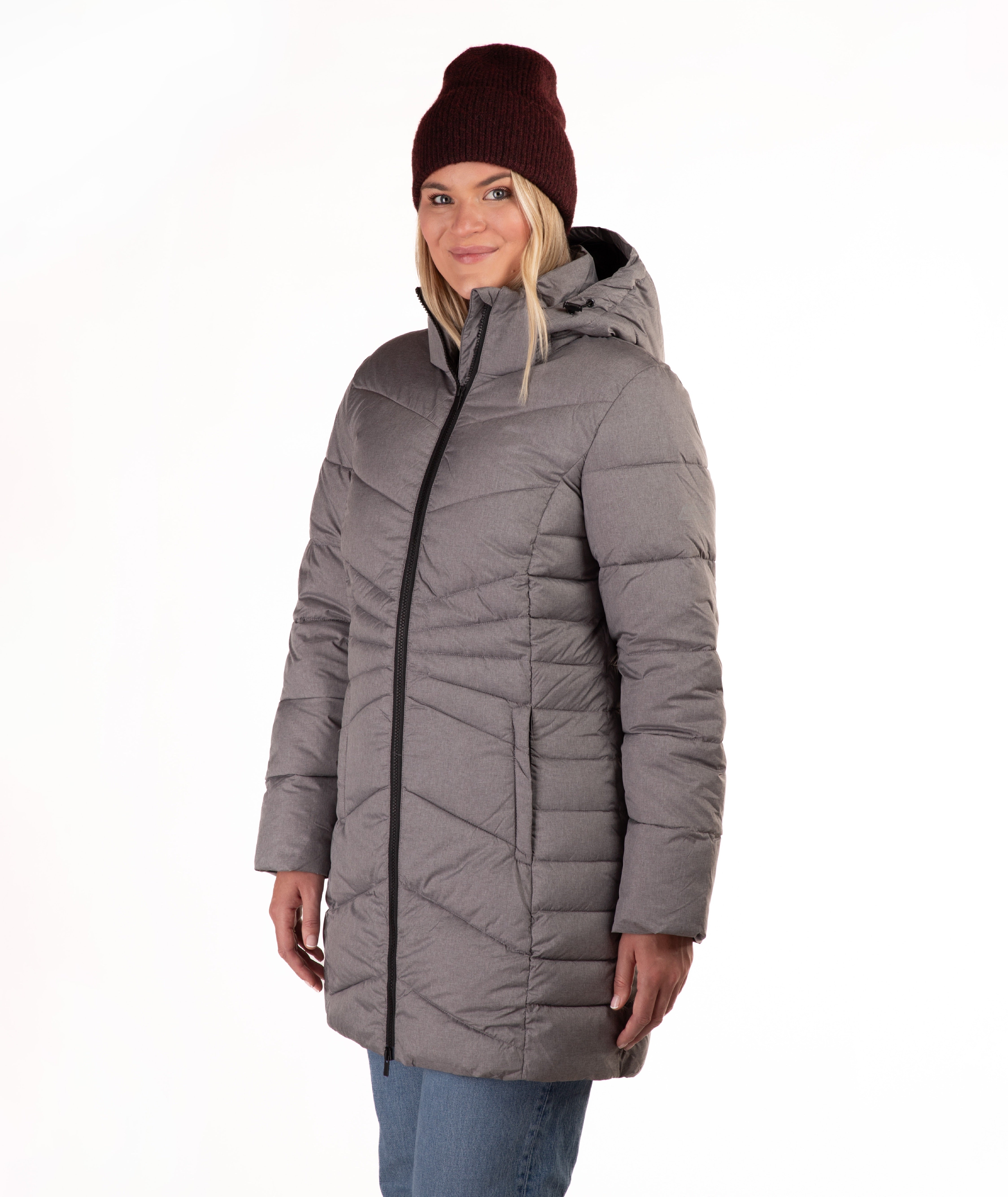 A woman wearing a gray hooded puffer jacket with a black front zipper, paired with a burgundy knit beanie and blue jeans, standing against a plain white background.