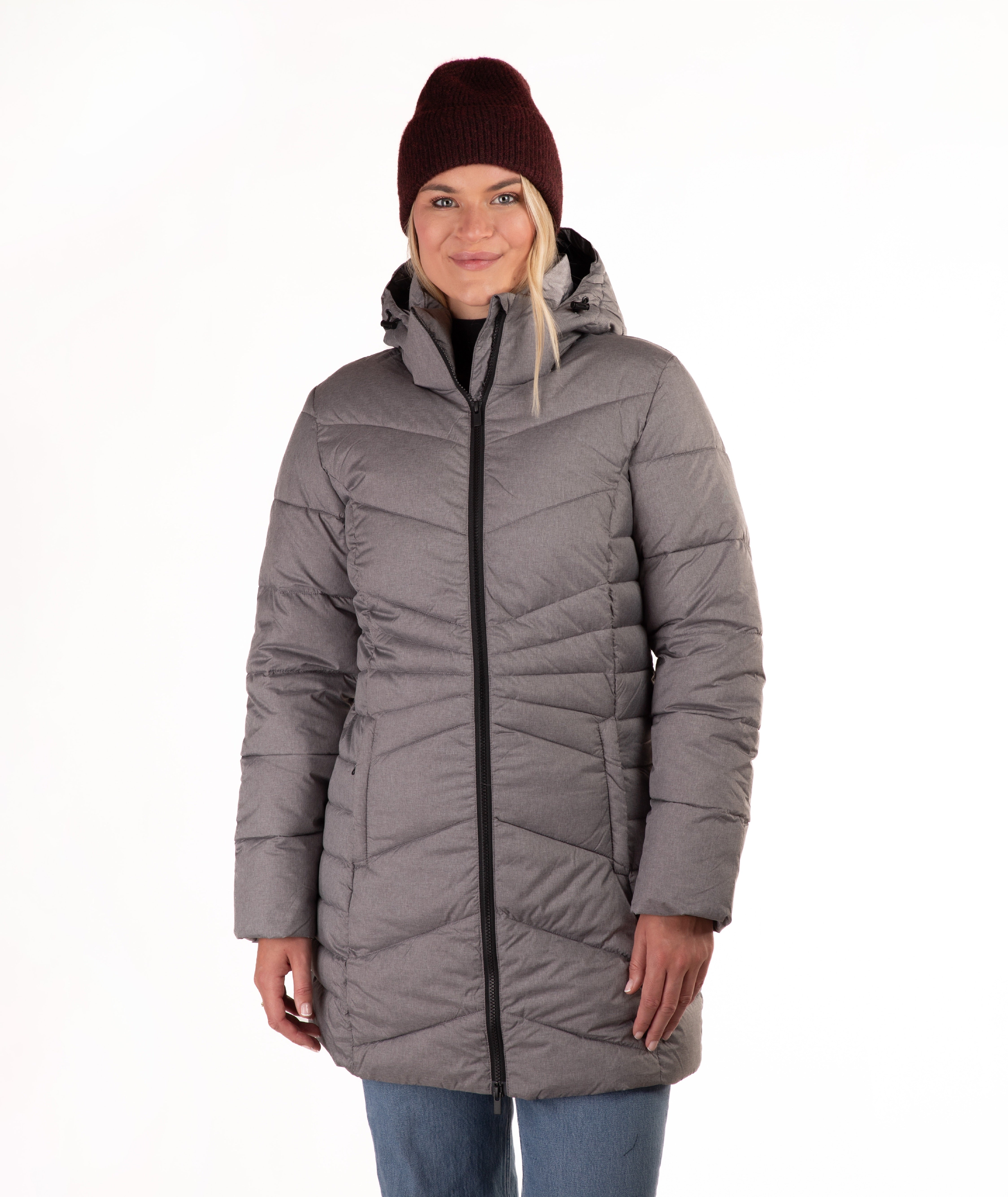 A woman wearing a gray hooded puffer jacket with a black front zipper, paired with a burgundy knit beanie and blue jeans, standing against a plain white background.