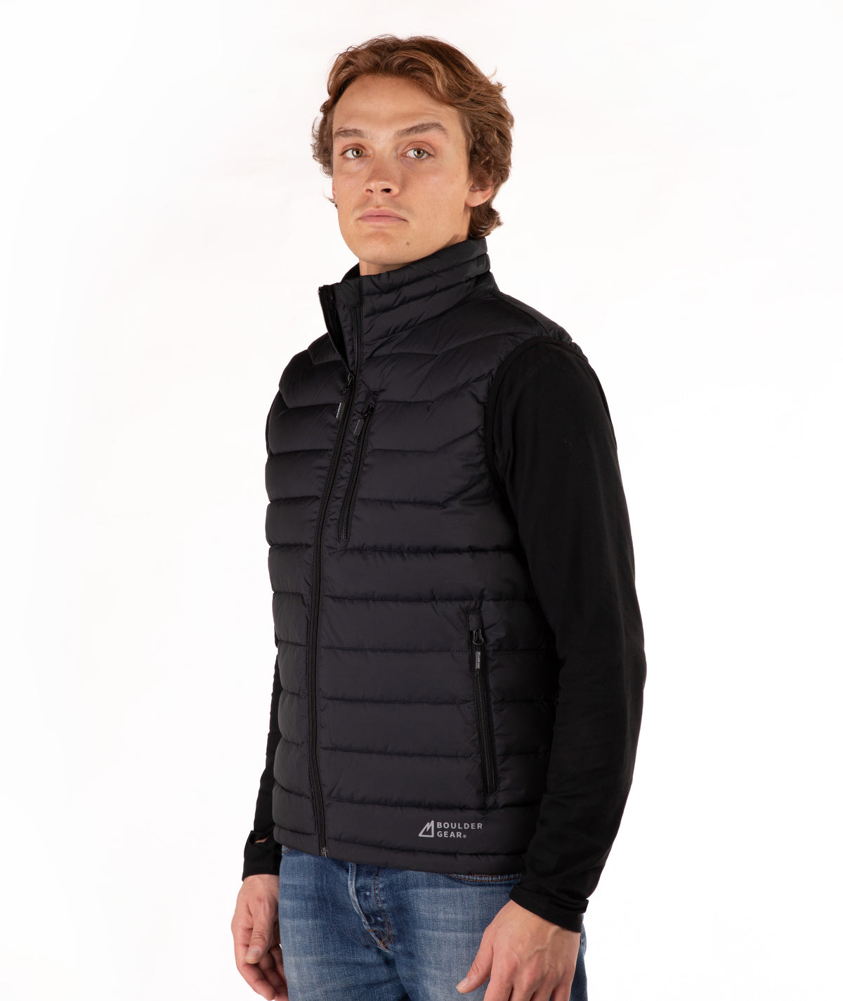 A man wearing a black quilted zip-up puffer vest over a black long-sleeve shirt and blue jeans, standing against a plain white studio background.