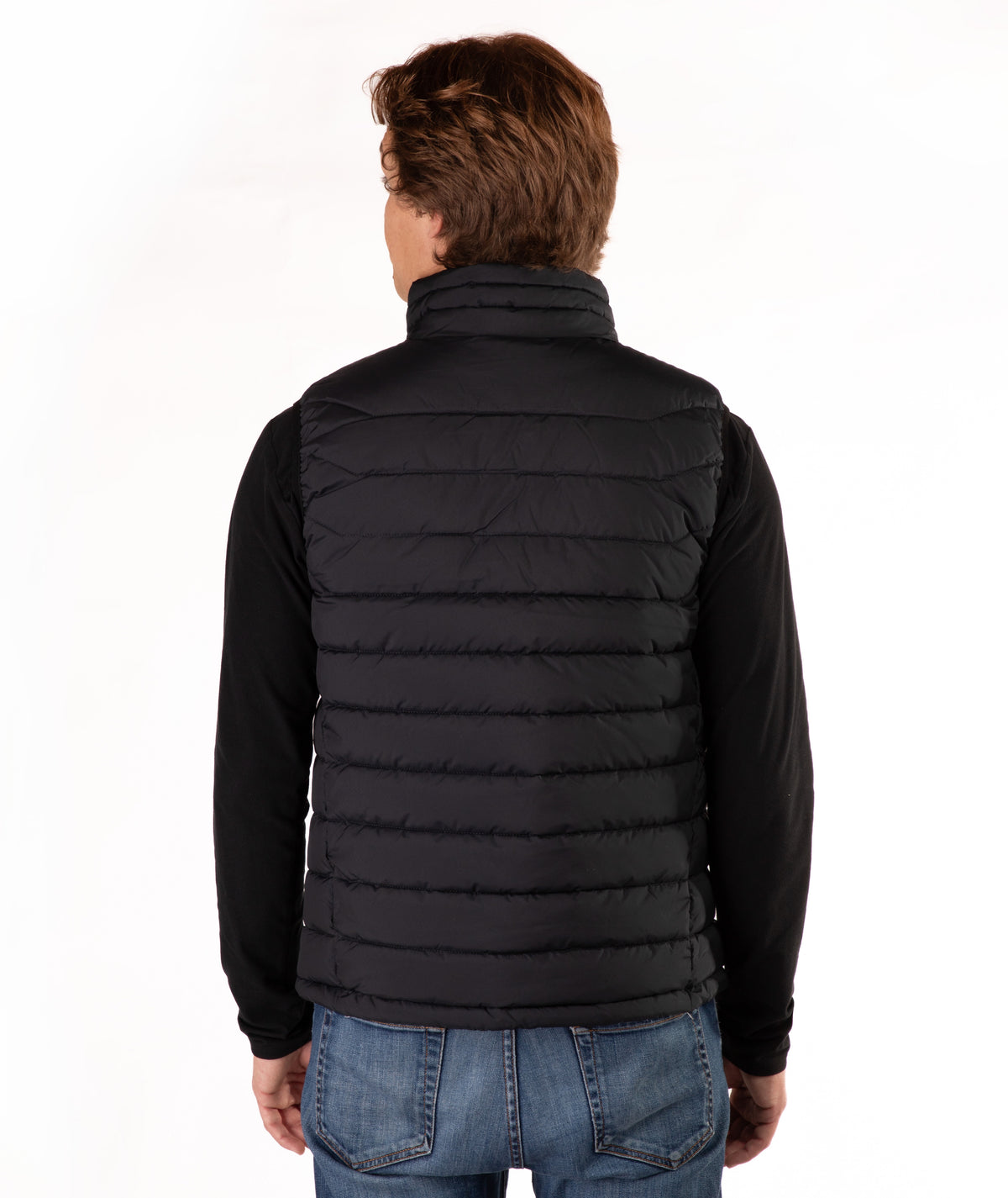 A man viewed from the back wearing a black quilted puffer vest over a black long-sleeve shirt and blue jeans, against a plain white studio background.