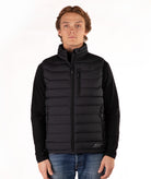 A man wearing a black quilted zip-up puffer vest over a black long-sleeve shirt and blue jeans, standing against a plain white studio background.