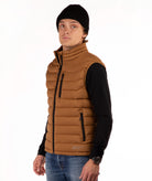 A man wearing a brown puffer vest over a black long-sleeve shirt, blue jeans, and a black beanie, standing against a plain white background.
