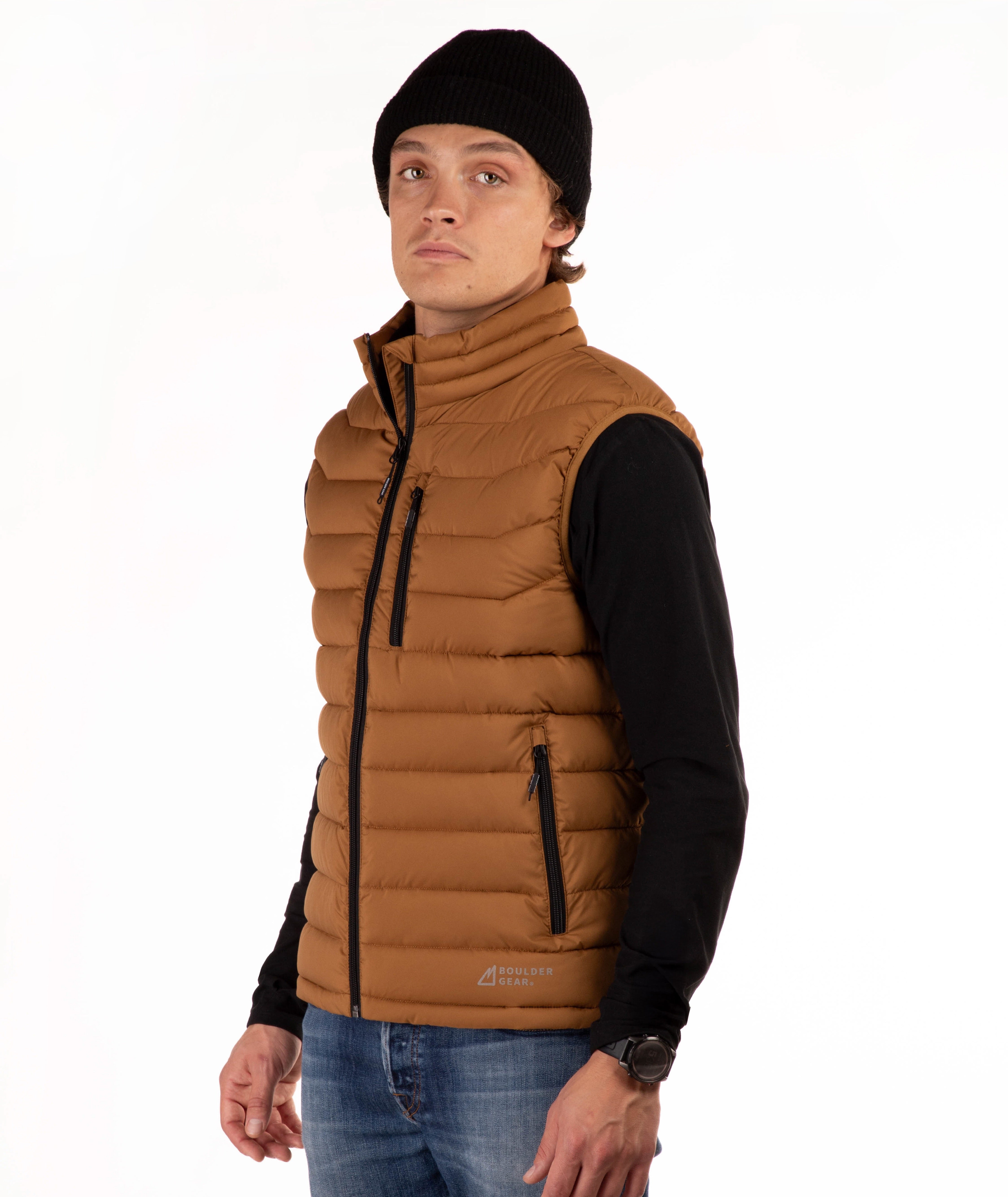 A man wearing a brown puffer vest over a black long-sleeve shirt, blue jeans, and a black beanie, standing against a plain white background.