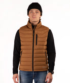 A man wearing a brown puffer vest over a black long-sleeve shirt, blue jeans, and a black beanie, standing against a plain white background.