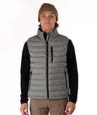 A man wearing a taupe puffer vest over a black long-sleeve shirt, with a brown beanie and khaki pants, standing against a plain white studio background.