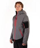 A man wearing a gray and black ski jacket with bright red zippers, standing against a plain white studio background.