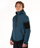 A man wearing a dark teal and black hooded technical jacket with a full front zipper, zippered hand pockets, and a black sleeve patch, photographed against a plain white studio background.