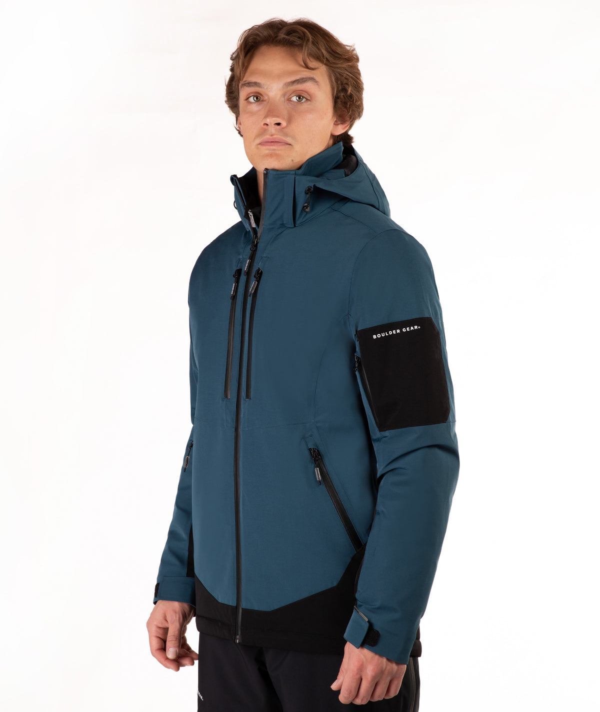 A man wearing a dark teal and black hooded technical jacket with a full front zipper, zippered hand pockets, and a black sleeve patch, photographed against a plain white studio background.