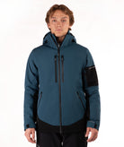 A man wearing a dark teal and black hooded technical jacket with a full front zipper, zippered hand pockets, and a black sleeve patch, photographed against a plain white studio background.