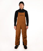 A male model wearing brown snow bib overalls with black panels, a black long-sleeve shirt, and a black beanie, standing against a solid white studio background.