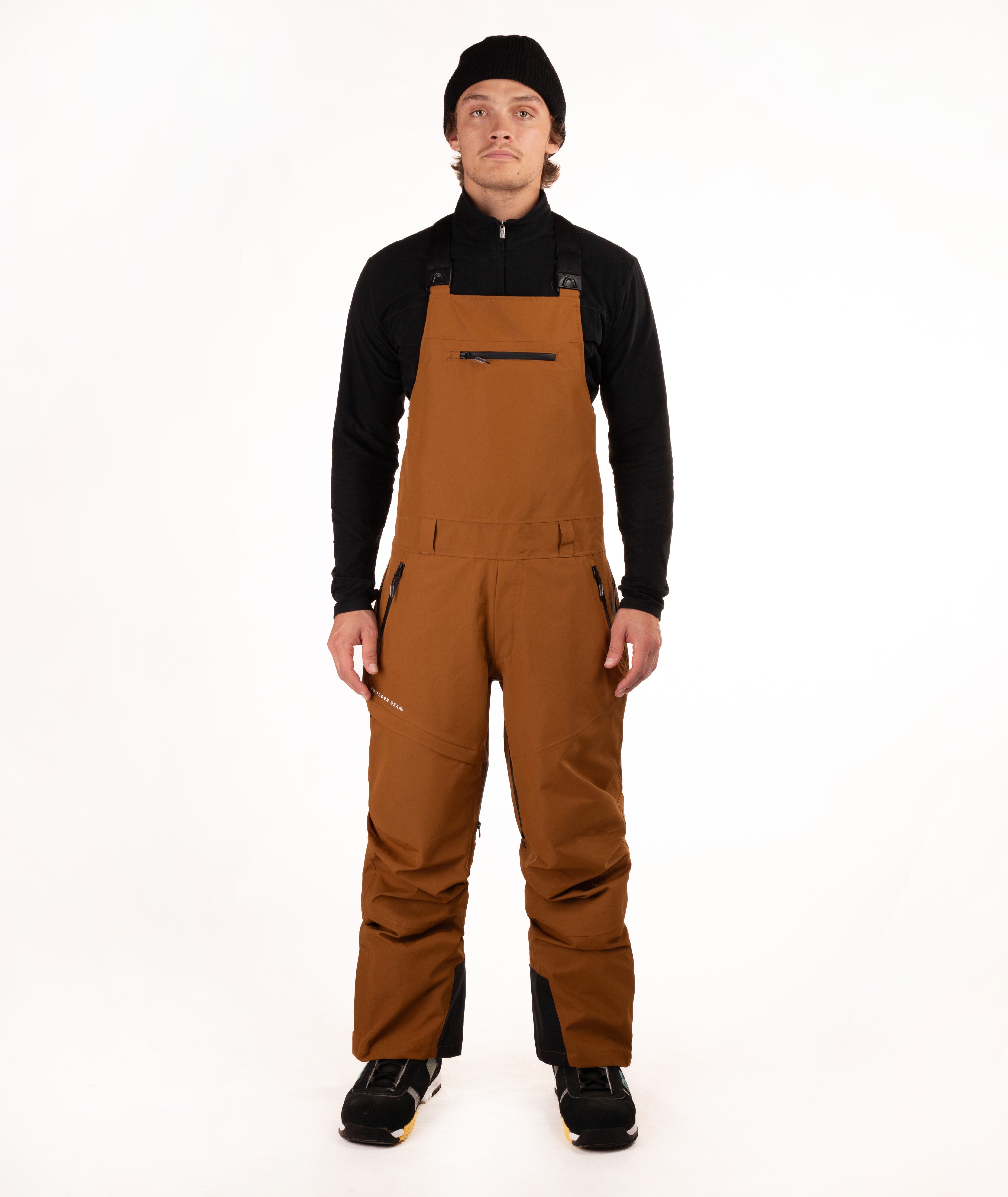 A male model wearing brown snow bib overalls with black panels, a black long-sleeve shirt, and a black beanie, standing against a solid white studio background.