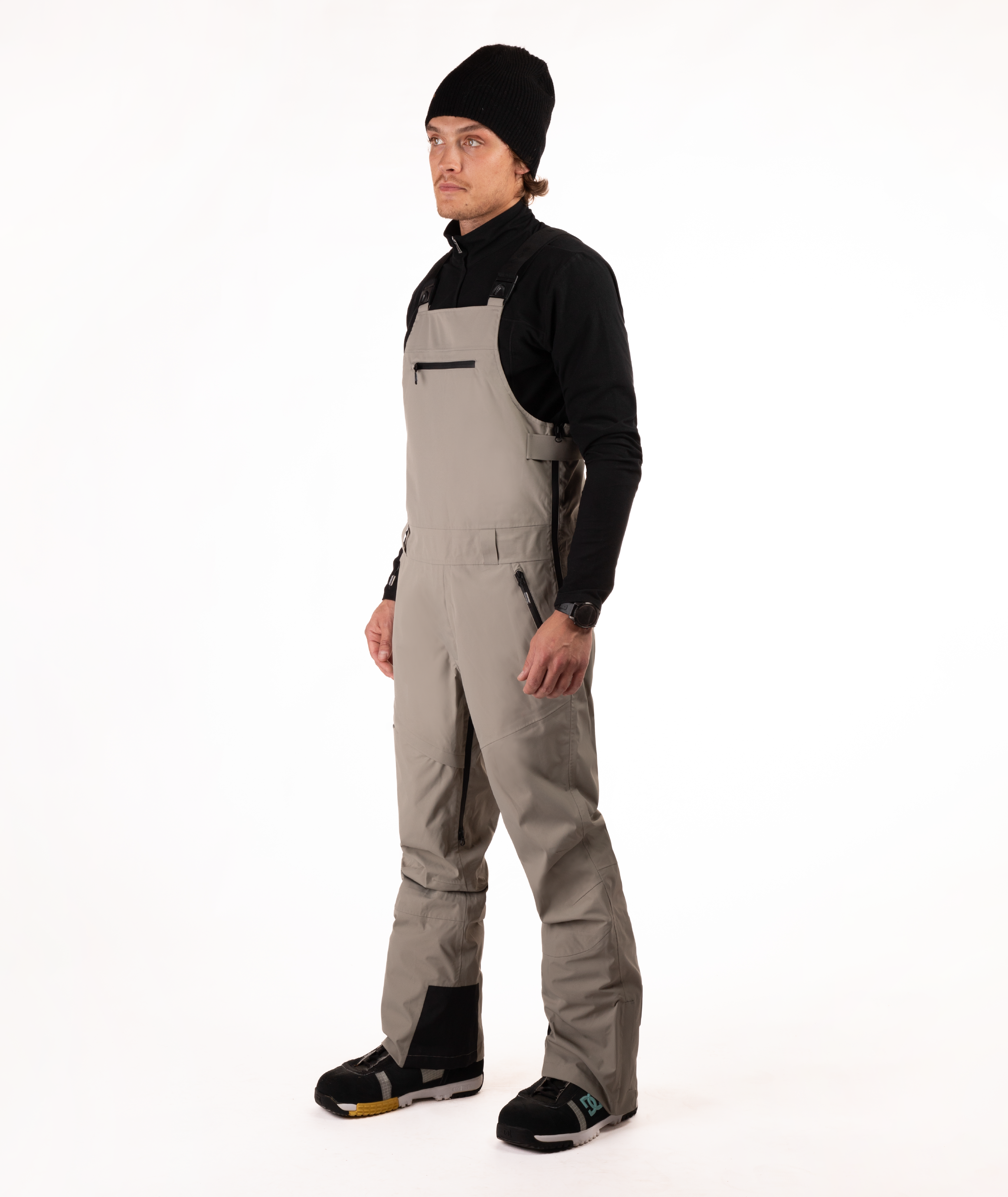 Person wearing a black long-sleeve shirt and beige bib overalls on a white background