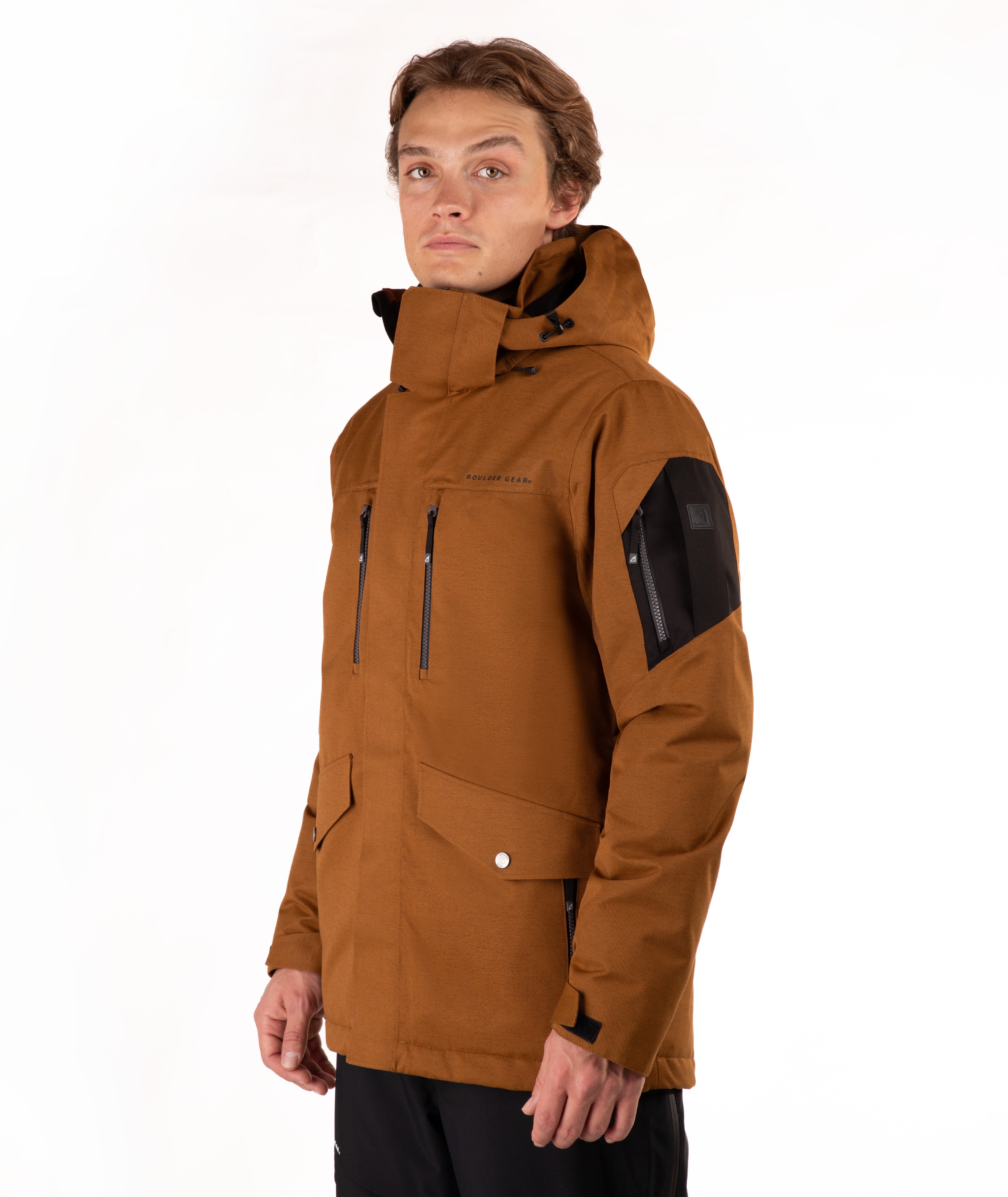 A man wearing a brown hooded technical jacket with black zippers, flap pockets, and a black sleeve patch, photographed against a plain white studio background.