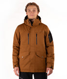 A man wearing a brown hooded technical jacket with black zippers, flap pockets, and a black sleeve patch, photographed against a plain white studio background.