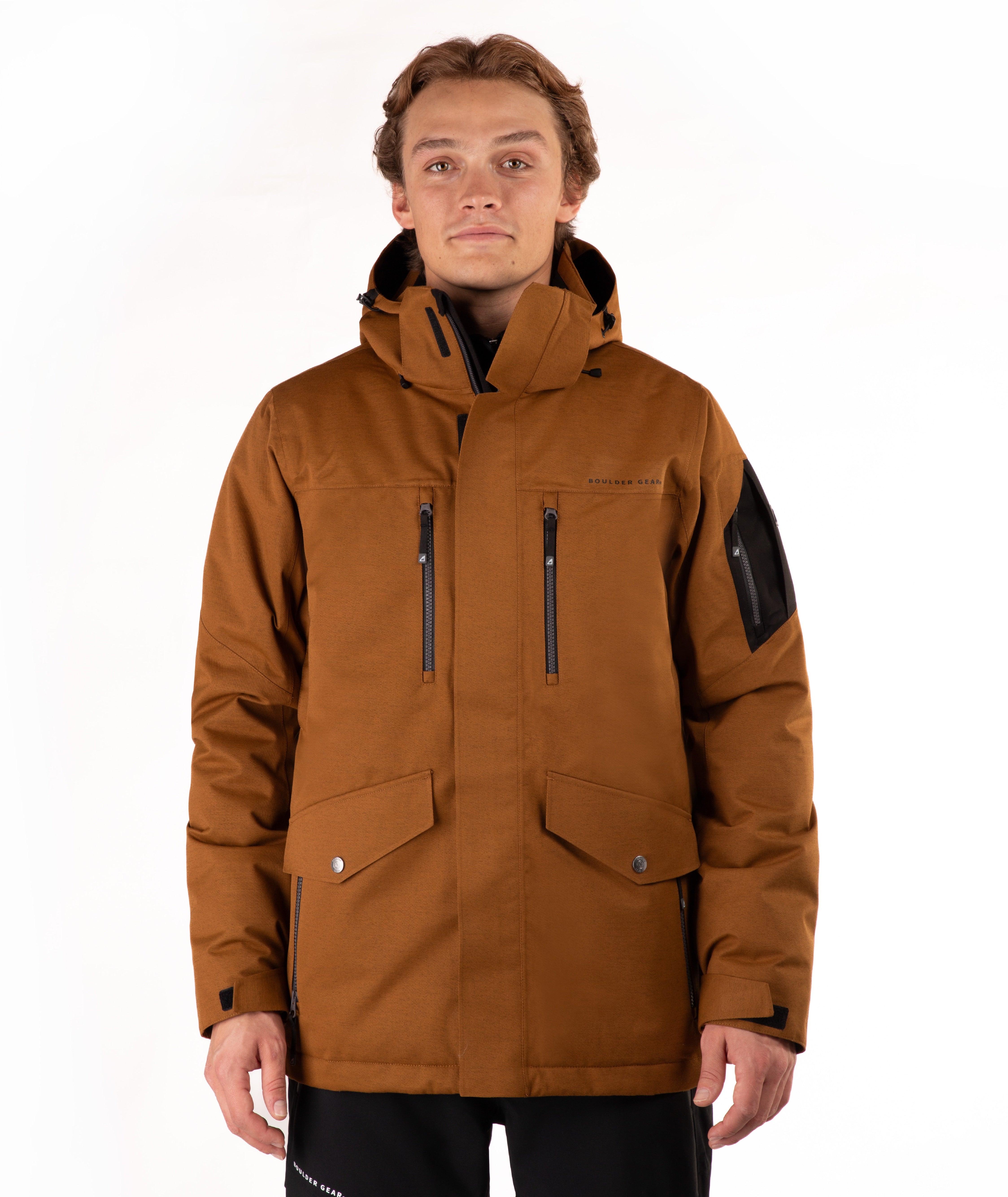 A man wearing a brown hooded technical jacket with black zippers, flap pockets, and a black sleeve patch, photographed against a plain white studio background.
