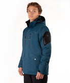 A man wearing a dark teal hooded outdoor jacket with black zippers, flap pockets, and a black sleeve patch, photographed against a plain white studio background.