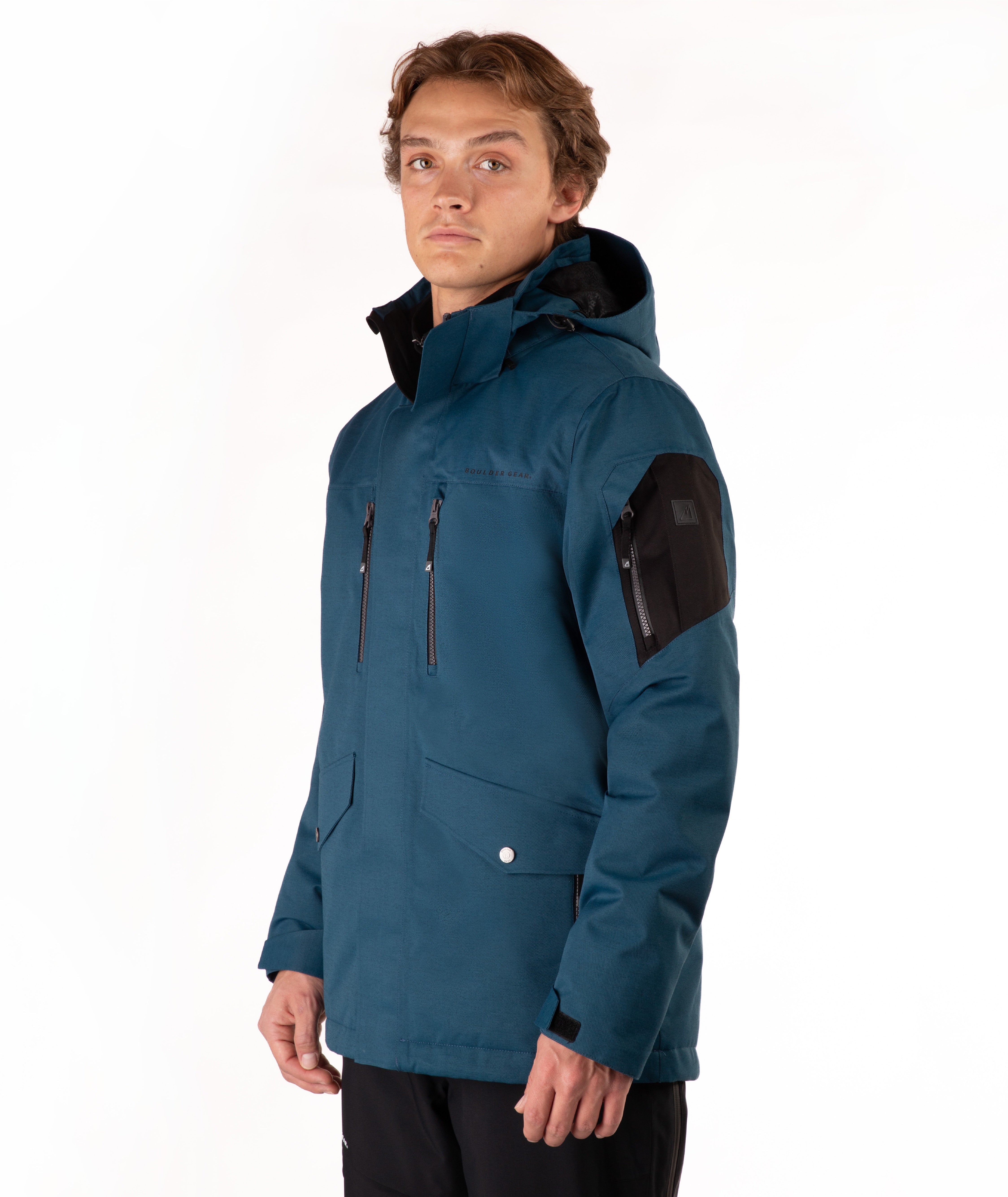 A man wearing a dark teal hooded outdoor jacket with black zippers, flap pockets, and a black sleeve patch, photographed against a plain white studio background.